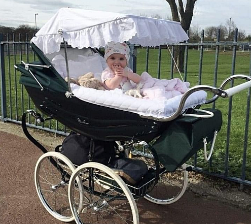  Klasyczny czarny wózek dziecięcy z białymi kołami i białym baldachimem z falbankami, stojący na ścieżce obok zielonego ogrodzenia. W wózku siedzi dziecko w różowej czapce i ubraniu, przykryte białym kocem, z pluszowym misiem obok. W tle widoczne drzewa i trawiaste pole.
